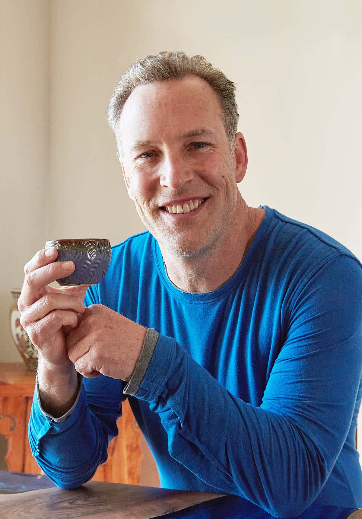 Lee Holden wearing a blue long sleeve t-shirt, smiling while holding a tea cup
