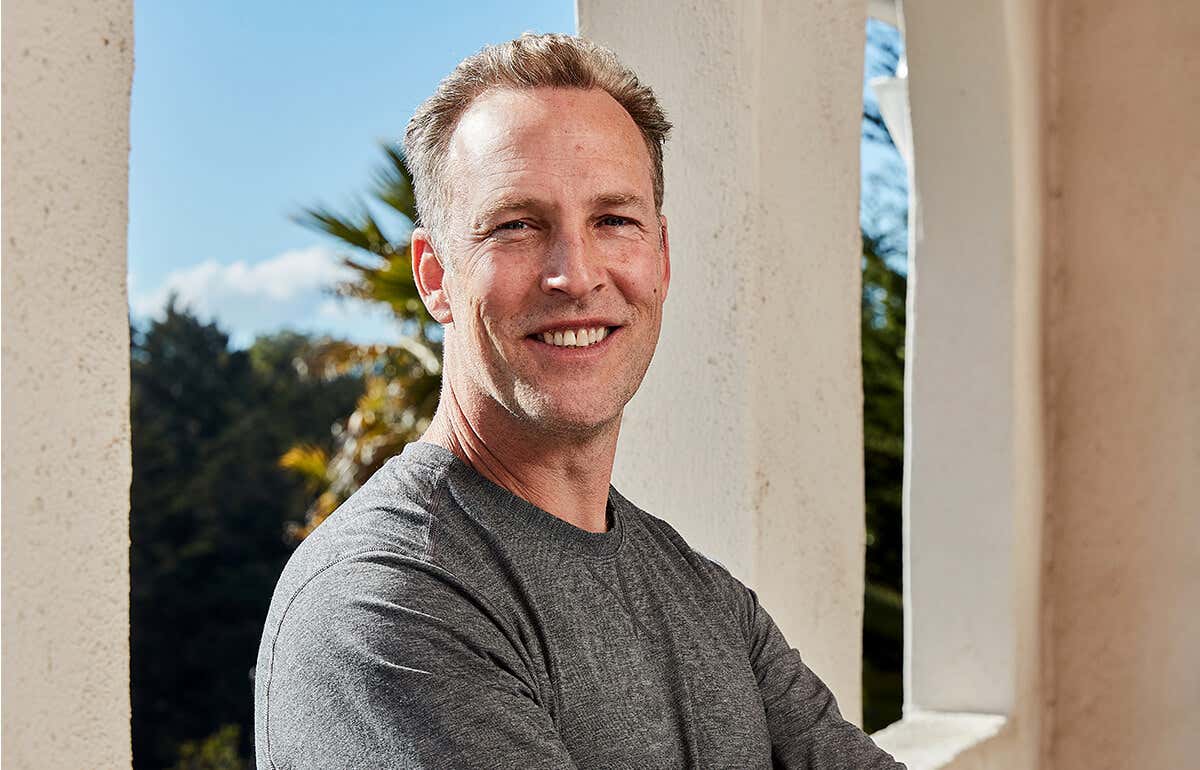 Lee Holden wearing a gray t-shirt, smiling with a balcony window backdrop