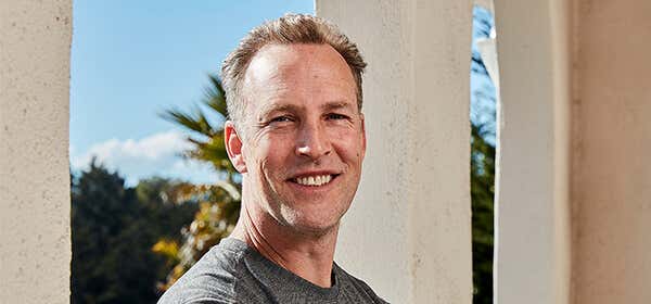 Lee Holden wearing a gray t-shirt, smiling with a balcony window backdrop