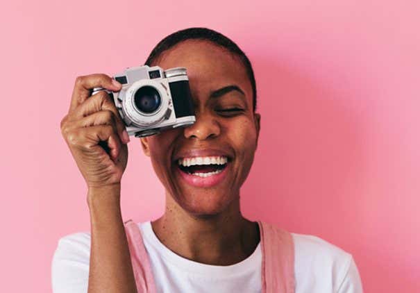 Young woman holding a vintage camera smiling in front of a pink background