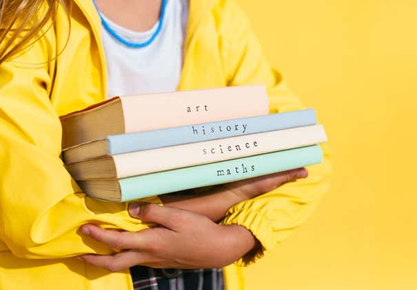 Girl in bright raincoat holding pile of various textbooks while standing against yellow background during studies at school