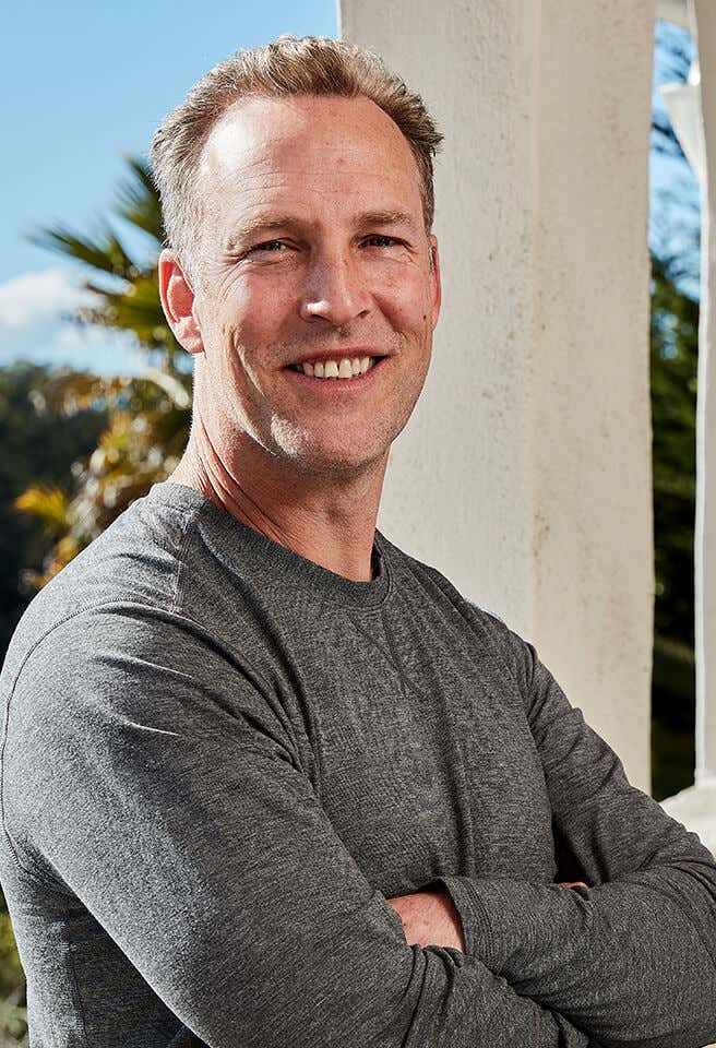 Lee Holden wearing a gray t-shirt, smiling with a balcony window backdrop