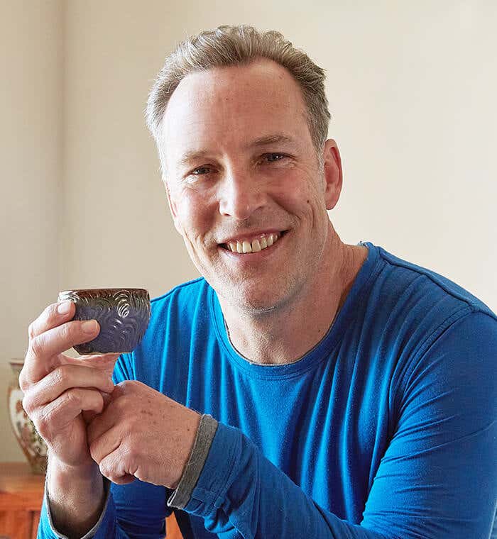 Lee Holden wearing a blue long sleeve t-shirt, smiling while holding a tea cup