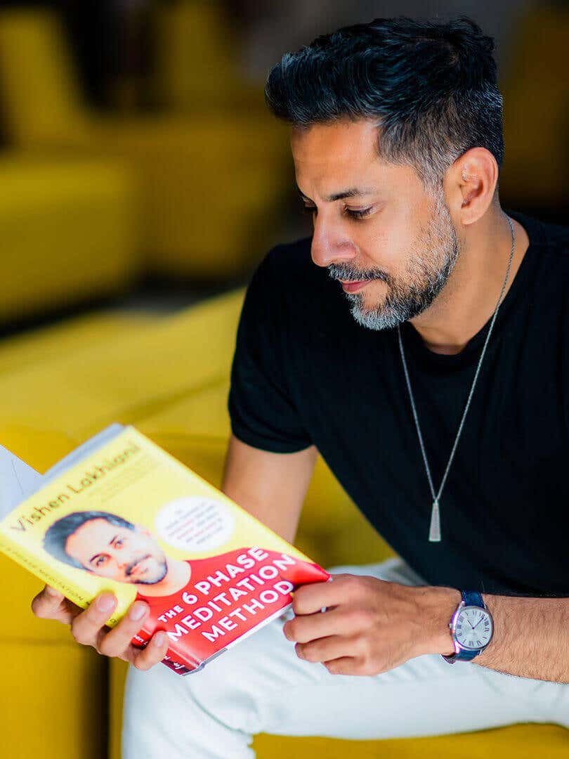 Vishen sitting on a yellow sofa, reading the 6 Phase Meditation Method book