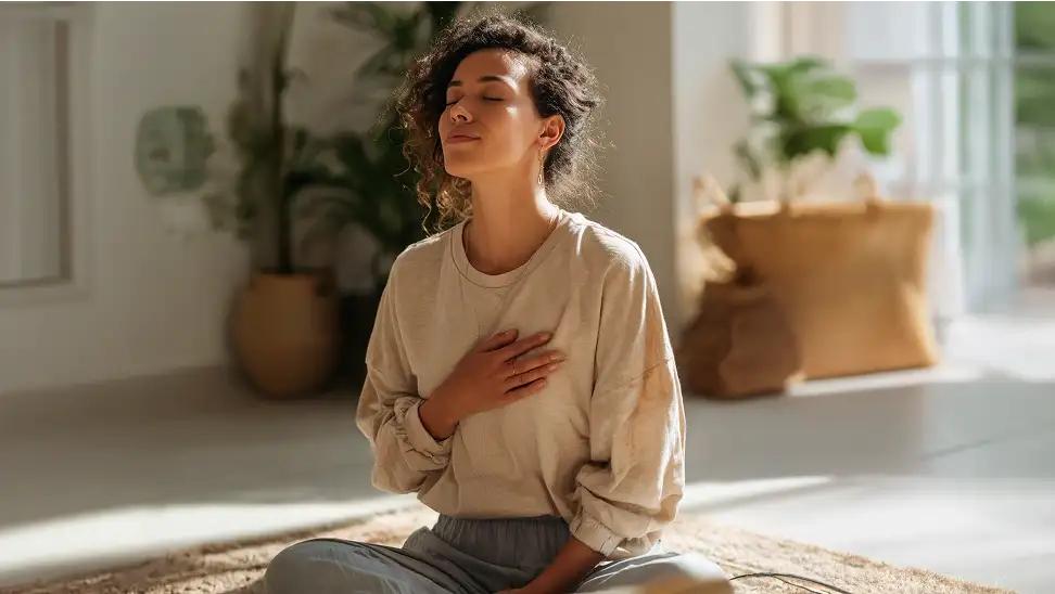 Woman practicing mindfulness meditation at home in a sunlit room with plants in the background.